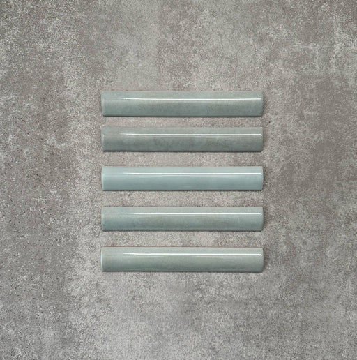 Flatlay mit Zellige Aquablau Bullnose-Keramikfliesen im marokkanischen Stil im Format 3 × 20 cm, die das abgerundete Kantenprofil, die glänzende blau-grüne Glasur und die feine tonale Variation vor neutralem Hintergrund zeigen.