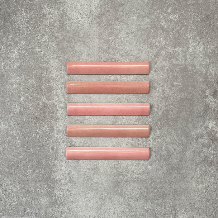 Flatlay mit Zellige Rosa Bordürenfliese im Format 3 × 20 cm im marokkanischen Stil. Keramische Abschlussfliesen in sanften Blush- und Pudertönen mit glatter, abgerundeter Kante und feiner Glasurvariation, arrangiert auf einem strukturierten grauen Hintergrund, um die handgefertigte Zellige-Optik und das dekorative Abschlussprofil hervorzuheben.