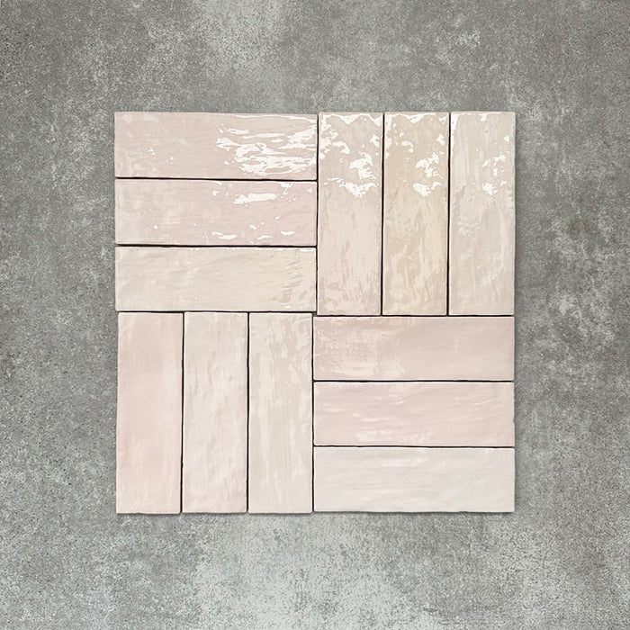 Flatlay von Zellige Rosa Nebel Ziegel Keramikfliesen im marokkanischen Stil im Format 6,5 × 20 cm, in sanften Rosa- und Blushtönen. Im Flechtmuster (Basketweave) auf strukturiertem grauem Hintergrund arrangiert, um die glänzende Glasur, die sanfte tonale Variation und die handgefertigte Zellige-Optik hervorzuheben.