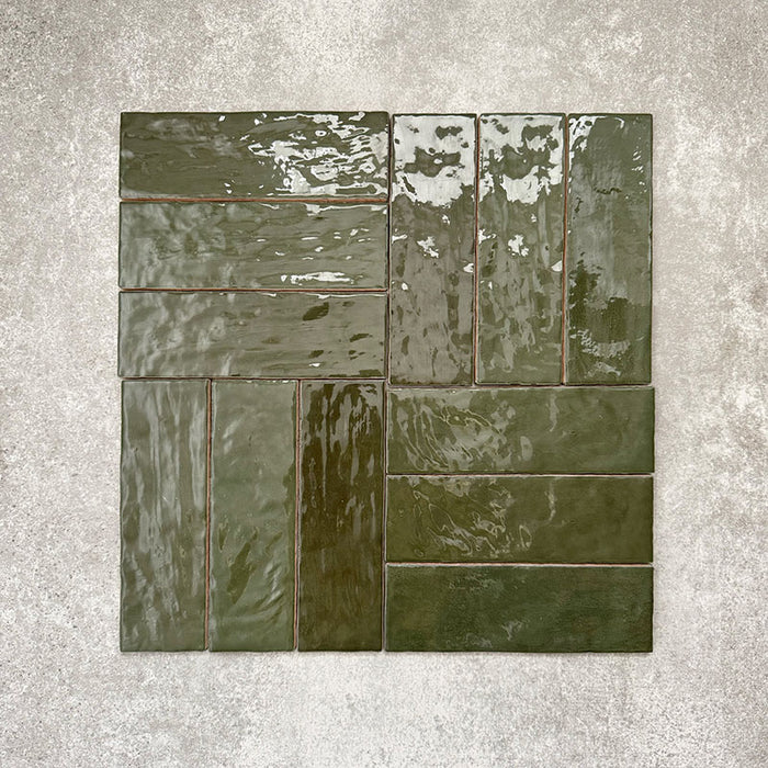Flatlay von Zellige Botanisches Grün Ziegel Keramikfliesen im marokkanischen Stil im Format 6,5 × 20 cm, im Flechtmuster (Basketweave) auf strukturiertem grauem Hintergrund angeordnet. Die glänzende Glasur, natürliche tonale Variation und die handgefertigte Zellige-Optik stehen im Fokus.