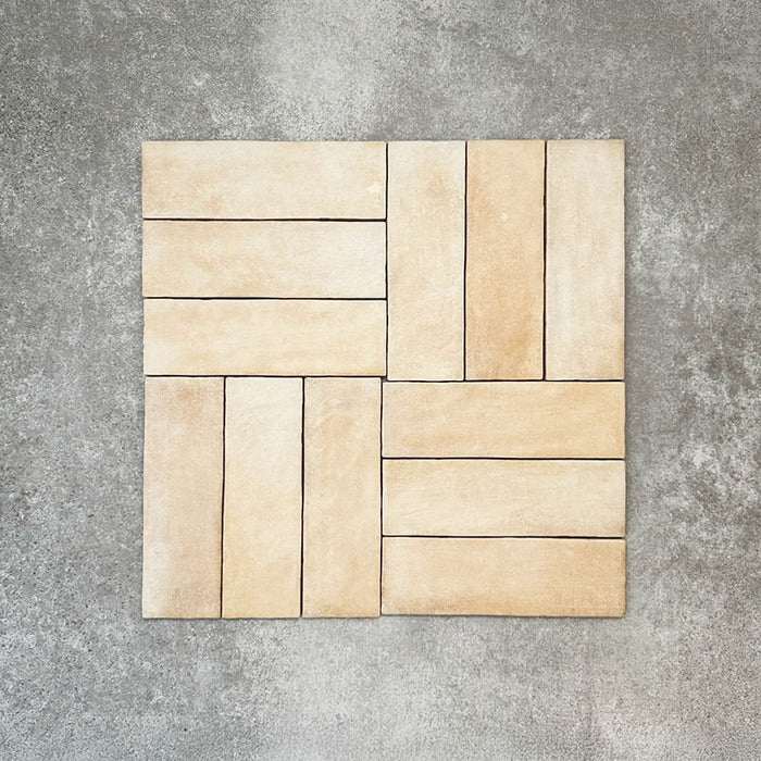 Flatlay von Zellige Sandbeige Matt Ziegel Keramikfliesen im marokkanischen Stil im Format 6,5 × 20 cm, in warmen Beige- und Cremetönen. Im Flechtmuster (Basketweave) auf strukturiertem grauem Hintergrund angeordnet, mit subtiler Farbvariation und leicht unregelmäßigen Kanten zur Betonung der handgefertigten Zellige-Optik.