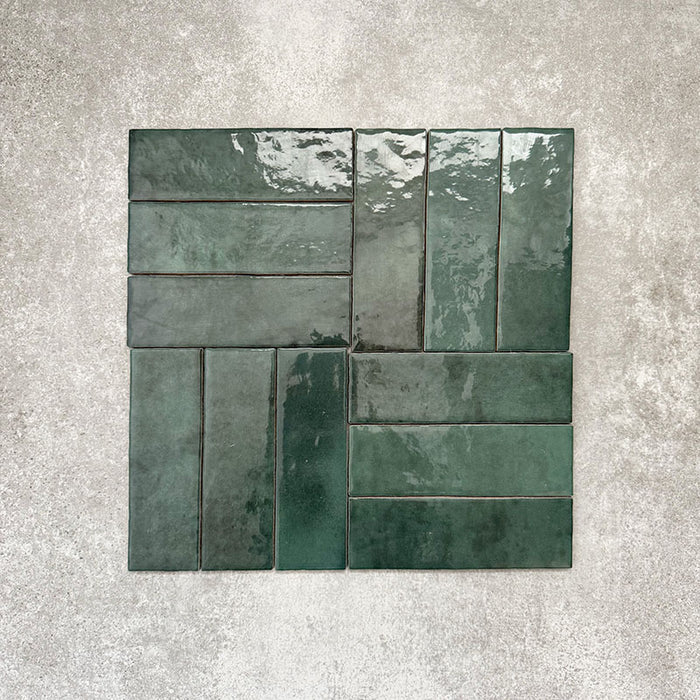Flatlay von Zellige Königliches Grün Ziegel Keramikfliesen im marokkanischen Stil im Format 6,5 × 20 cm, im Flechtmuster (Basketweave) auf strukturiertem grauem Hintergrund arrangiert. Die glänzende Glasur, natürliche Farbvariation und die handgefertigte Zellige-Optik stehen im Fokus.