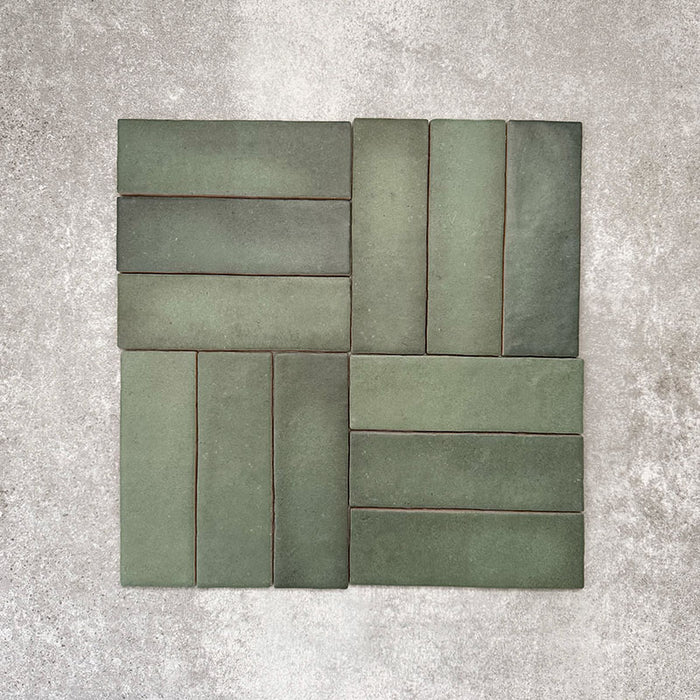 Flatlay von Zellige Malachitgrün Matt Keramik-Riemchenfliesen im marokkanischen Stil im Format 6,5 × 20 cm, im Flechtmuster (Basketweave) arrangiert. Sichtbar sind die feine Farbvariation, die handgefertigte Textur und der satte Grünton vor neutralem Hintergrund.