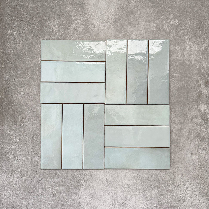 Flatlay von Zellige Aquablau Keramik-Riemchenfliesen im marokkanischen Stil im Format 6,5 × 20 cm, im Flechtmuster (Basketweave) angeordnet. Sanfte Blau-Grün-Töne, glänzende Glasur in Handarbeit-Optik und feine Farbvariation werden vor neutralem Hintergrund präsentiert.