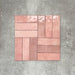 Flatlay von Zellige Rosa Brick Keramikfliesen im marokkanischen Stil im Format 6,5 × 20 cm, in sanften Blush-, Altrosa- und Terrakotta-Pinktönen mit feiner Farbvariation und leicht unregelmäßigen Kanten. Im Flechtmuster (Basketweave) auf strukturiertem grauem Hintergrund arrangiert, um die handwerkliche Zellige-Optik zu zeigen.