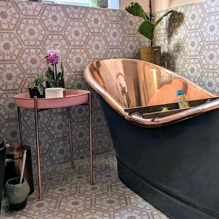 Badezimmer mit Medina Rosa Feinsteinzeug-Hexagonfliesen im marokkanischen Stil im Format 18,7 × 21,6 cm, an Wand und Boden verlegt. Das sanfte geometrische Muster in Rosatönen bildet einen ruhigen Hintergrund und wird mit einer Badewanne in Kupferoptik sowie Vintage-Accessoires stilvoll ergänzt.