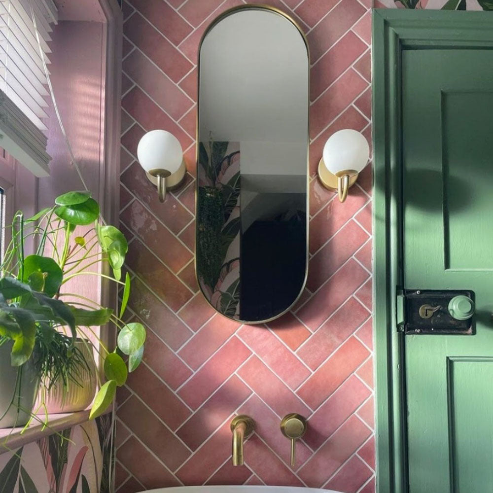 Badezimmerwand mit Zellige Rosa Brick Keramikfliesen im marokkanischen Stil im Format 6,5 × 20 cm, im Fischgrätmuster verlegt. Sanfte Blush- und Pudertöne in Rosa werden mit Wand- und Armaturen in Messing, einer grün gestrichenen Tür und einem Spiegel mit Rundbogen kombiniert – für ein warmes, charaktervolles Finish mit handgefertigter Zellige-Optik.