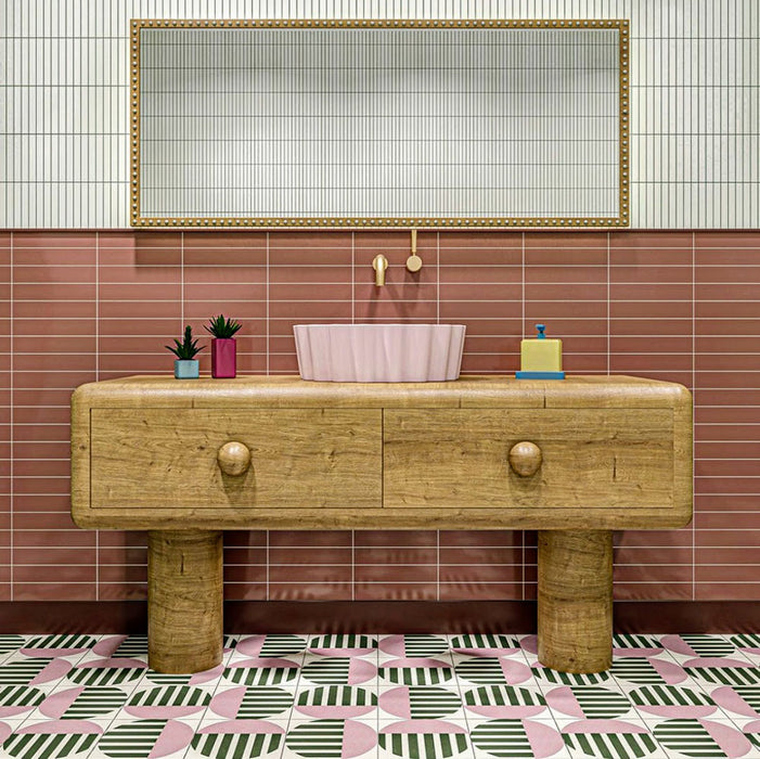 Badezimmerinterieur mit Abstrakter Serit Musterfliese im Format 20 × 20 cm, verlegt als markantes geometrisches Bodendesign. Das sich wiederholende Muster kombiniert sanfte Rosatöne, tiefes Grün und klares Weiß mit gestreiften Details und wird durch einen Waschtisch aus Holz sowie ein rosafarbenes Waschbecken ergänzt – für ein verspieltes und zugleich zeitgemäßes Badezimmerambiente.