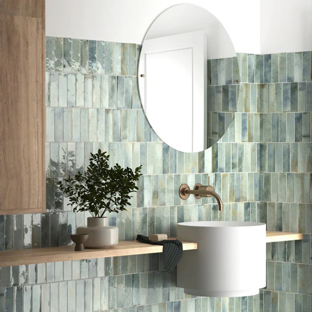 Badezimmerwand mit Saigon Seladongrün Brick Wandfliesen im marokkanisch inspirierten Stil. Glänzende Keramikfliesen im Format 5,1 × 16,1 cm in sanften Grün- und Blautönen, kombiniert mit einem runden Spiegel und einem minimalistischen Waschtisch.
