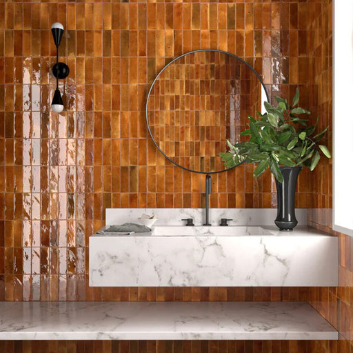 Badezimmerwand mit Saigon Bernstein Brick Wandfliesen im marokkanisch inspirierten Stil. Glänzende Keramikfliesen im Format 5,1 × 16,1 cm in warmen Bernsteintönen, kombiniert mit einem Waschtisch in Marmoroptik und einem runden Spiegel.