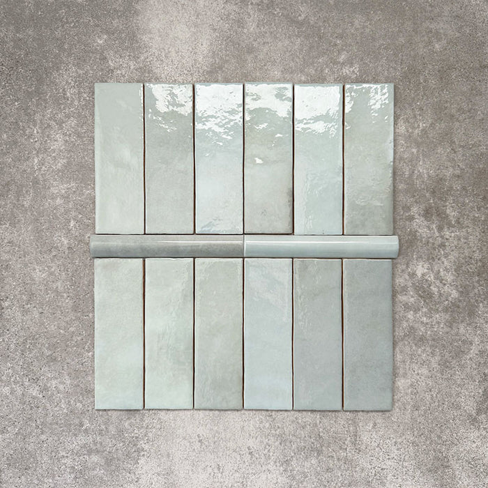Flatlay mit Zellige Aquablau Keramikfliesen im marokkanischen Stil, bestehend aus 6,5 × 20 cm Riemchenfliesen in Kombination mit einer 3 × 20 cm Bullnose-Abschlussfliese. Das Arrangement hebt das abgerundete Kantenprofil, die glänzende blau-grüne Glasur und die natürliche Farbvariation vor neutralem Hintergrund hervor.