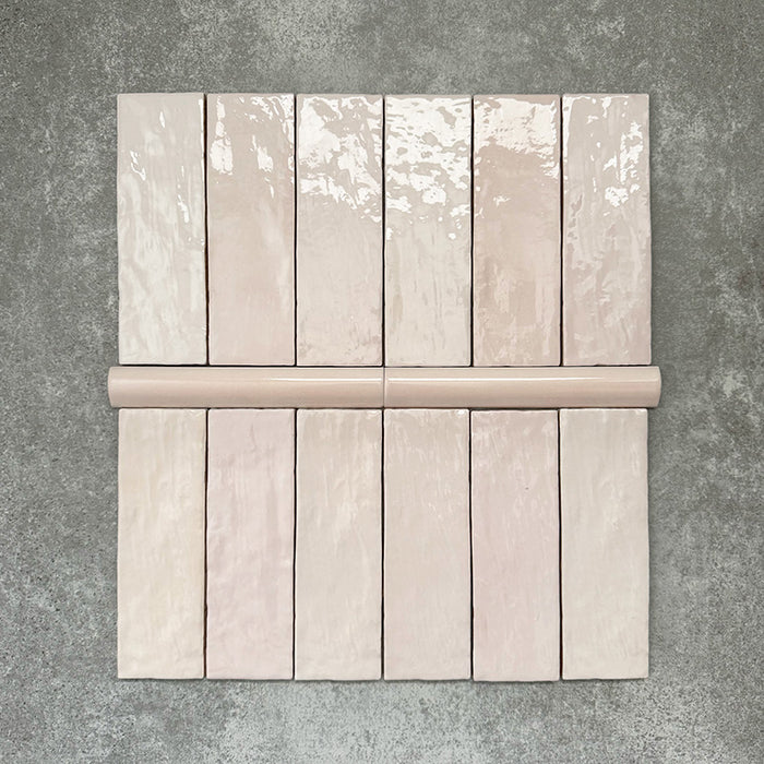 Flatlay mit Zellige Rosa Nebel Brick Keramikfliesen im marokkanischen Stil im Format 6,5 × 20 cm, kombiniert mit einer passenden Pink Mist Bullnose-Abschlussfliese im Format 3 × 20 cm. In sanften Rosétönen auf einem strukturierten grauen Hintergrund angeordnet, werden die glänzende Glasur, die feine tonale Variation und die handgefertigte Zellige-Optik hervorgehoben.