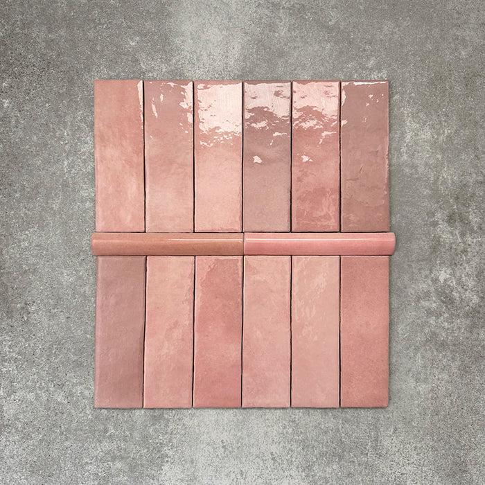 Flatlay mit Zellige Rosa Brick Keramikfliesen im marokkanischen Stil im Format 6,5 × 20 cm, kombiniert mit einer passenden rosafarbenen Bullnose-Abschlussfliese im Format 3 × 20 cm. Sanfte Blush- und Pudertöne mit natürlicher Glasurvariation und leicht unregelmäßigen Kanten, arrangiert auf einem strukturierten grauen Hintergrund, zeigen das abgestimmte Zusammenspiel von Riemchenfliese und abgerundetem Abschlussdetail für eine handgefertigte Zellige-Optik.
