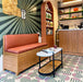Café-Interieur mit Abstrakter Serit Musterfliese im Format 20 × 20 cm, verlegt als markantes geometrisches Bodendesign. Das verspielte Muster kombiniert sanft rosafarbene Halbkreise mit tiefgrünen Streifendetails auf weißem Grund und wird durch warme Holzmöbel und rosafarbene Sitzmöbel ergänzt – für einen zeitgemäßen, charaktervollen Gewerberaum.