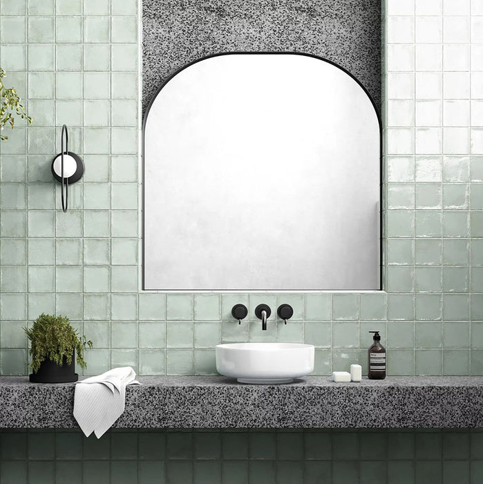 Badezimmer mit Dordogne Matcha Green Rustic Gloss Wandfliesen im Format 10 × 10 cm in sanften Grüntönen mit dezenten Farbvariationen und rustikaler Glanzoberfläche, kombiniert mit Terrazzo-Oberflächen, schwarzen Armaturen und einem bogenförmigen Spiegel für einen modernen Look.