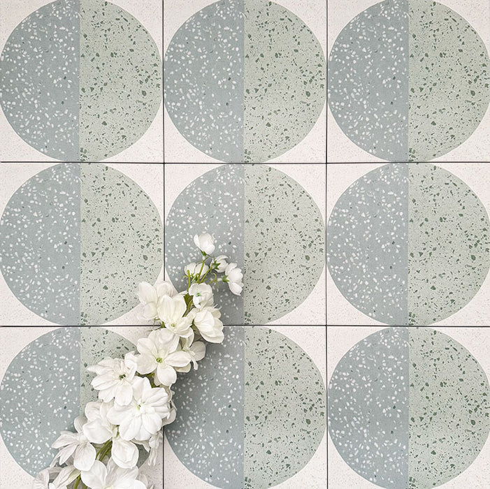 Close-up of Eclipse Salvia 20x20cm terrazzo-effect decorative tiles in soft sage and grey tones, featuring geometric circular patterns with speckled detailing, styled with white floral accents.