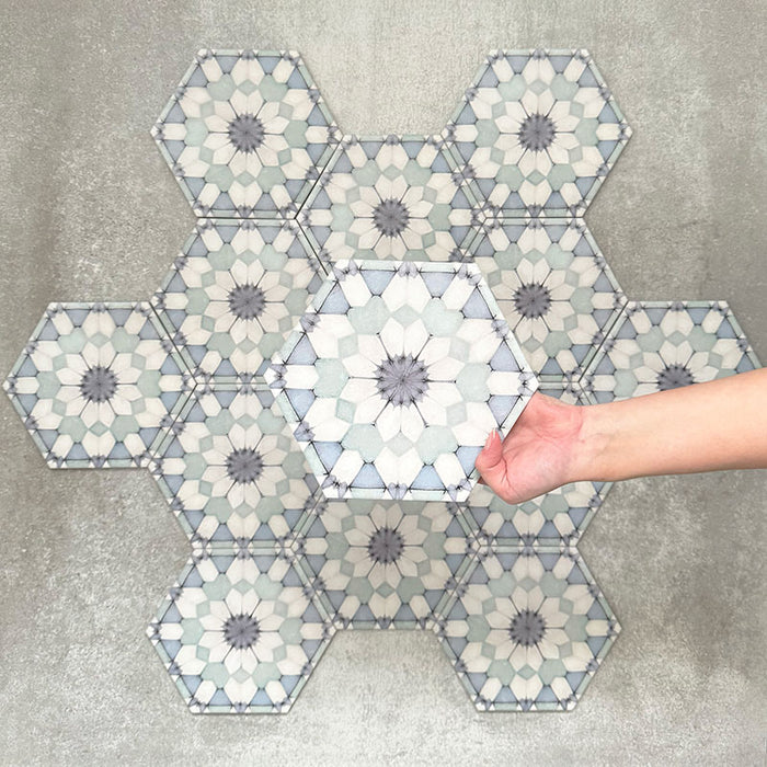 Hand hält eine Medina Frostblau Feinsteinzeug-Hexagonfliese im marokkanischen Stil im Format 18,7 × 21,6 cm, gezeigt vor einer gruppierten Anordnung, um das sich wiederholende geometrische Muster in sanften Blau- und Weißtönen hervorzuheben.