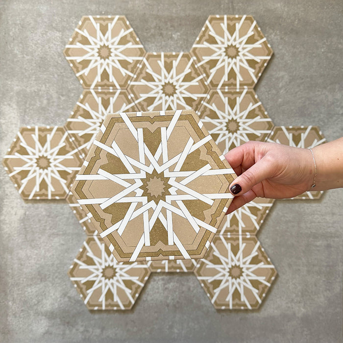 Hand hält eine Medina Hormuz Vintage Gold Hexagon-Porzellanfliese im marokkanischen Stil, 18,7 × 21,6 cm, vor einer gruppierten Anordnung, um das markante sterninspirierten geometrische Muster in warmen Gold- und Weißtönen hervorzuheben.