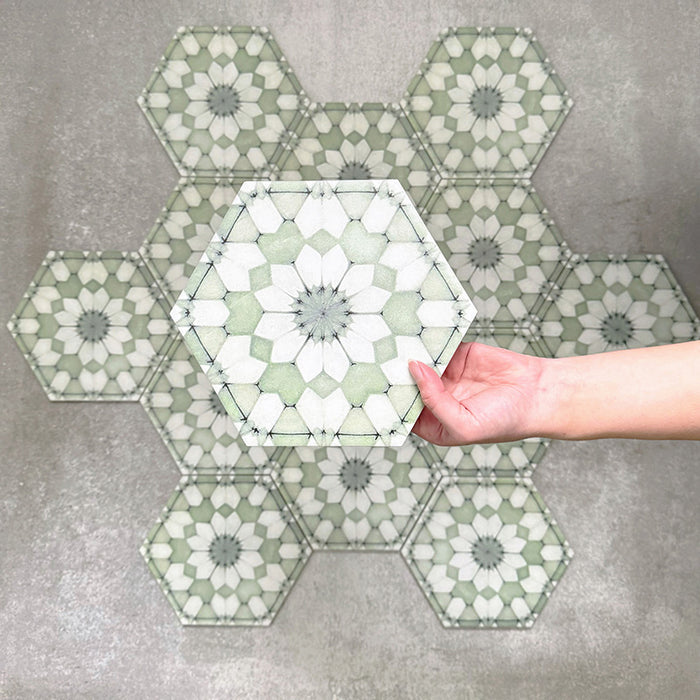 Hand hält eine Medina Soft Grün Feinsteinzeug-Hexagonfliese im marokkanischen Stil im Format 18,7 × 21,6 cm. Dargestellt vor einer gruppierten Anordnung, um das sich wiederholende geometrische Muster und die sanften Grüntöne hervorzuheben.
