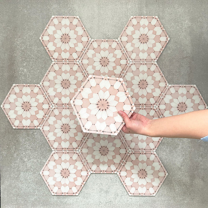 Hand hält eine Medina Rosa Feinsteinzeug-Hexagonfliese im marokkanischen Stil im Format 18,7 × 21,6 cm, dargestellt vor einer gruppierten Anordnung, um das sich wiederholende geometrische Muster in sanften Rosa- und Weißtönen hervorzuheben.