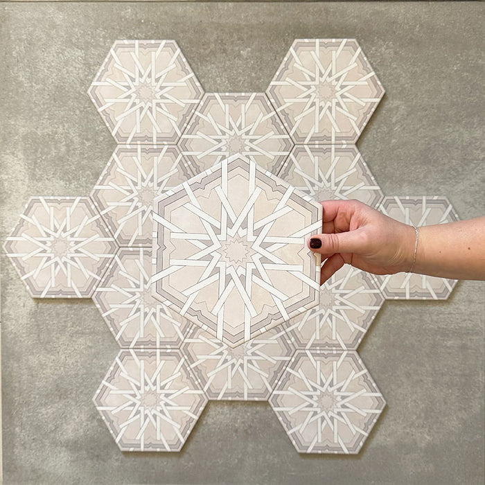 Hand hält eine Medina Hormuz Schneeweiß Feinsteinzeug-Hexagonfliese im marokkanischen Stil im Format 18,7 × 21,6 cm, dargestellt vor einer gruppierten Anordnung, um das klare, sterninspirierte geometrische Muster in Weiß- und hellen Grautönen hervorzuheben.