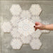 Hand hält eine Medina Hormuz Schneeweiß Feinsteinzeug-Hexagonfliese im marokkanischen Stil im Format 18,7 × 21,6 cm, dargestellt vor einer gruppierten Anordnung, um das klare, sterninspirierte geometrische Muster in Weiß- und hellen Grautönen hervorzuheben.