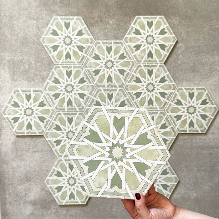 Hand hält eine Medina Hormuz Grün Feinsteinzeug-Hexagonfliese im marokkanischen Stil im Format 18,7 × 21,6 cm, dargestellt vor einer gruppierten Anordnung, um das kräftige Sternmuster in Grün- und Weißtönen hervorzuheben.