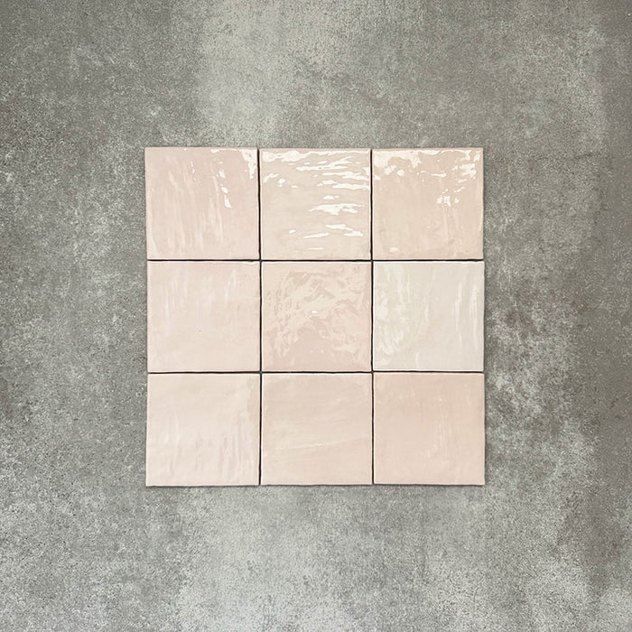 Flatlay von Zellige Rosa Nebel Quadrat Keramikfliesen im marokkanischen Stil im Format 13,2 × 13,2 cm, in sanften Rosa- und Blushtönen. Im einfachen Raster auf strukturiertem grauem Hintergrund angeordnet, um die glänzende Glasur, die dezente Farbvariation und die handgefertigte Zellige-Optik zu zeigen.