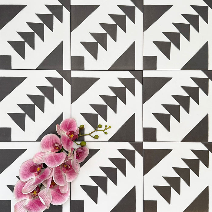 Almeira Azteca Feinsteinzeugfliesen im marokkanischen Stil, angeordnet in einem sich wiederholenden Muster, das das markante geometrische Design mit gestuften Dreiecken hervorhebt. Mit rosafarbenen Blumen kombiniert, um einen spannenden Kontrast zu schaffen, Format 20 × 20 cm.