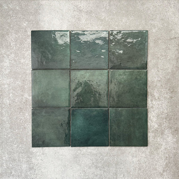 Flatlay von Zellige Königliches Grün Quadrat Keramikfliesen im marokkanischen Stil im Format 13,2 × 13,2 cm, in satten, tiefen Grüntönen. Im einfachen Raster auf strukturiertem grauem Hintergrund angeordnet, um die glänzende Glasur, feine tonale Variation und die handgefertigte Zellige-Optik zu zeigen.