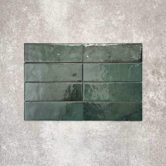 Flatlay von Zellige Königliches Grün Ziegel Keramikfliesen im marokkanischen Stil im Format 6,5 × 20 cm, horizontal im Ziegelverband auf strukturiertem grauem Hintergrund angeordnet, um die glänzende Glasur, natürliche Farbvariation und den handgefertigten Charakter zu zeigen.