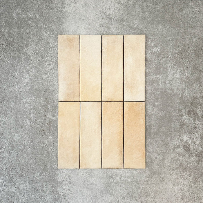 Flatlay von Zellige Sandbeige Matt Ziegel Keramikfliesen im marokkanischen Stil im Format 6,5 × 20 cm, in einer warmen Beige- und Cremefarbpalette. Mit feiner tonaler Variation und sanft unregelmäßigen Kanten, vertikal im Ziegelverband auf strukturiertem grauem Hintergrund arrangiert, um den handgefertigten Zellige-Effekt hervorzuheben.
