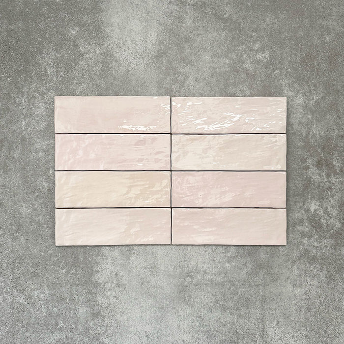 Flatlay von Zellige Rosa Nebel Ziegel Keramikfliesen im marokkanischen Stil im Format 6,5 × 20 cm, in sanften Rosa- und Blushtönen. Horizontal im Ziegelverband auf strukturiertem grauem Hintergrund arrangiert, um die glänzende Glasur, die sanfte tonale Variation und die handgefertigte Zellige-Optik zu präsentieren.