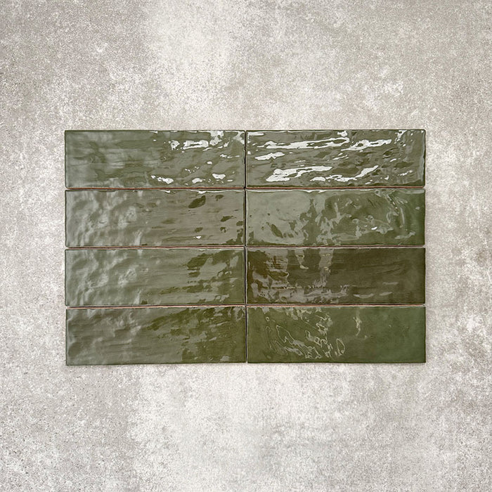 Flatlay von Zellige Botanisches Grün Ziegel Keramikfliesen im marokkanischen Stil im Format 6,5 × 20 cm, horizontal im Ziegelverband auf strukturiertem grauem Hintergrund verlegt, um die glänzende Glasur, die feine Farbvariation und die handgefertigte Zellige-Optik hervorzuheben.
