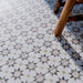 Marketingmotiv mit Agadir Beige Fliesen im marokkanischen Stil im Format 20 × 20 cm aus Feinsteinzeug, verlegt als Bodenbelag mit einem sanften, neutralen geometrischen Sternmuster in dezenten Grau- und warmen Beigetönen. Mit einem Holzstuhl in Szene gesetzt, um eine wohnliche, authentische Atmosphäre zu vermitteln.
