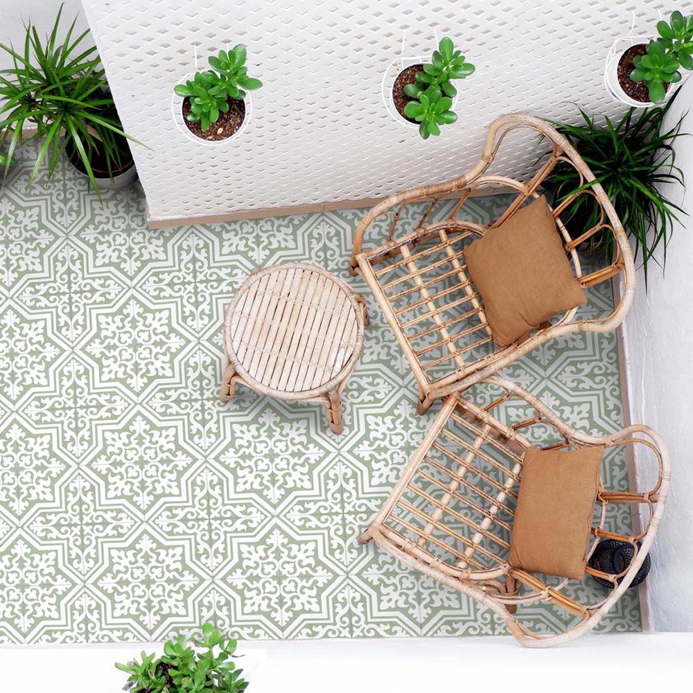 Außenbereich mit Sitzplatz, ausgestattet mit Rattanstühlen, einem kleinen runden Tisch und hängenden Pflanzen, gestaltet mit Berlin Trondite Grün Bodenfliesen im Format 20 × 20 cm im viktorianisch-marokkanisch inspirierten Stil. Gezeigt wird ein frisches Muster in Salbei und Weiß.