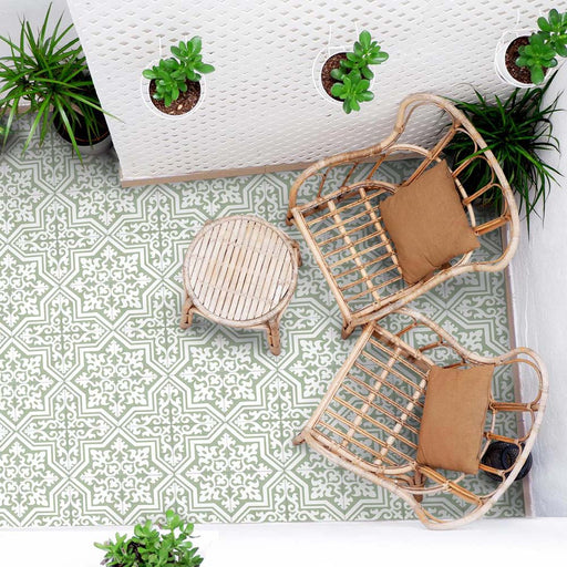 Außenbereich mit Sitzplatz, ausgestattet mit Rattanstühlen, einem kleinen runden Tisch und hängenden Pflanzen, gestaltet mit Berlin Trondite Grün Bodenfliesen im Format 20 × 20 cm im viktorianisch-marokkanisch inspirierten Stil. Gezeigt wird ein frisches Muster in Salbei und Weiß.
