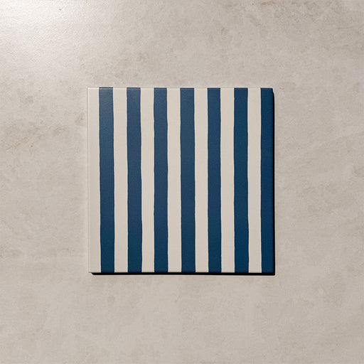 Produktpräsentation der Gestreift Blau Dekorfliesen im Format 20 × 20 cm in sanften Blau- und Weißtönen, mit unregelmäßigen vertikalen Streifen in handgemalter Optik vor einem Hintergrund in dezenter Steinoptik.