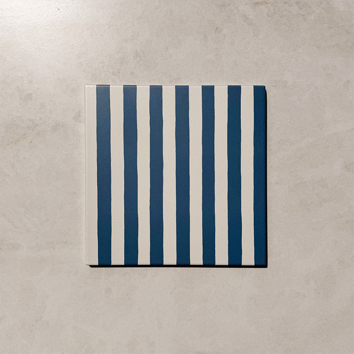 Produktpräsentation der Gestreift Blau Dekorfliesen im Format 20 × 20 cm in sanften Blau- und Weißtönen, mit unregelmäßigen vertikalen Streifen in handgemalter Optik vor einem Hintergrund in dezenter Steinoptik.