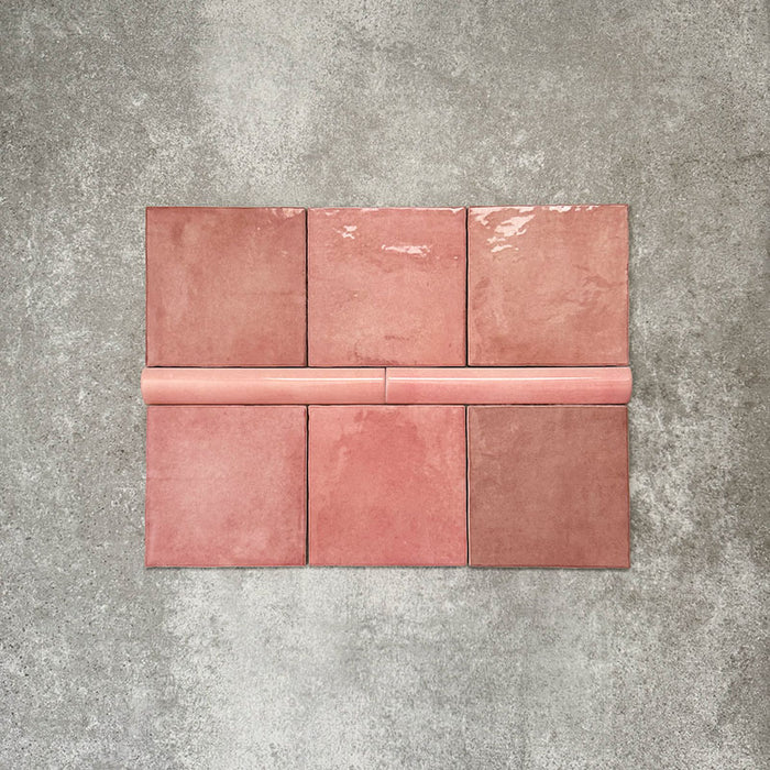 Flatlay mit Zellige Rosa Quadrat Keramikfliesen im marokkanischen Stil im Format 13,2 × 13,2 cm, kombiniert mit einer passenden rosafarbenen Bullnose-Abschlussfliese im Format 3 × 20 cm. Sanfte Blush- und Pudertöne mit natürlicher Glasurvariation und leicht unregelmäßigen Kanten, arrangiert auf einem strukturierten grauen Hintergrund, verdeutlichen das harmonische Zusammenspiel von Quadratfliese und abgerundetem Abschlussdetail für eine handgefertigte Zellige-Optik.