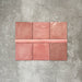Flatlay mit Zellige Rosa Quadrat Keramikfliesen im marokkanischen Stil im Format 13,2 × 13,2 cm, kombiniert mit einer passenden rosafarbenen Bullnose-Abschlussfliese im Format 3 × 20 cm. Sanfte Blush- und Pudertöne mit natürlicher Glasurvariation und leicht unregelmäßigen Kanten, arrangiert auf einem strukturierten grauen Hintergrund, verdeutlichen das harmonische Zusammenspiel von Quadratfliese und abgerundetem Abschlussdetail für eine handgefertigte Zellige-Optik.