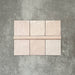 Flatlay mit Zellige Rosa Nebel Quadrat Keramikfliesen im marokkanischen Stil im Format 13,2 × 13,2 cm, kombiniert mit einer passenden Pink Mist Bullnose-Abschlussfliese im Format 3 × 20 cm. Sanfte, helle Rosétöne, natürliche Glasurvariation und leicht unregelmäßige Kanten werden auf einem strukturierten grauen Hintergrund gezeigt und unterstreichen die handgefertigte Zellige-Optik.