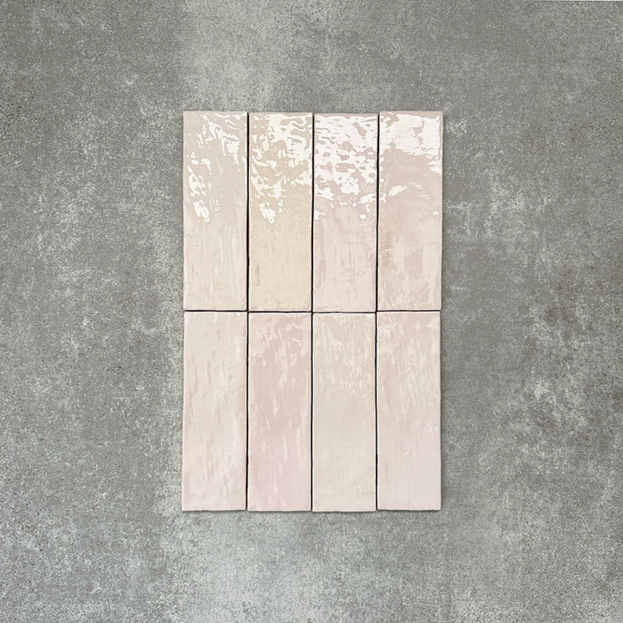 Flatlay von Zellige Rosa Nebel Ziegel Keramikfliesen im marokkanischen Stil im Format 6,5 × 20 cm, in zarten Rosa- und Blushtönen. Vertikal im Ziegelverband auf strukturiertem grauem Hintergrund angeordnet, um die glänzende Glasur, die sanfte Farbvariation und die handgefertigte Zellige-Optik zu zeigen.