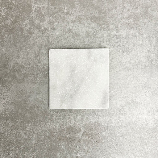 Einzelne Fayrouz White 15,2 × 15,2 cm Naturmarmorfliese mit getrommelter Oberfläche in sanften Weißtönen mit dezenter natürlicher Maserung, präsentiert vor einem neutralen, strukturierten Hintergrund in einer klaren Produktszene.