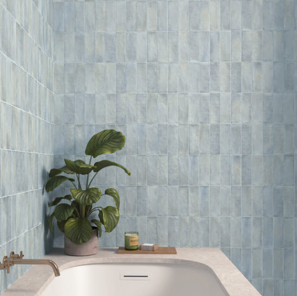Badezimmer mit Zellige Sky Blue Matt Brick-Wandfliesen im Format 6,5 × 20 cm im Moroccan Style, in sanften Himmelblautönen mit dezenten Farbvariationen und matter Oberfläche. Die Fliesen schaffen eine beruhigende Kulisse rund um eine moderne Badewanne und werden durch warme Armaturen in Messingoptik sowie natürliche Pflanzen ergänzt.